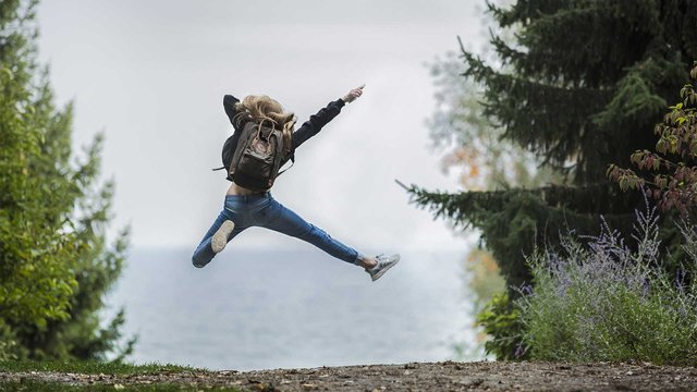 A woman jumping with a rucksack in the countryside, a lake in the background.